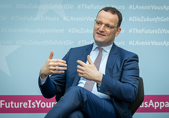 Bundesminister Jens Spahn zurückgelehnt auf einem Stuhl sitzend, etwas mit den Händen gestikulierend. Im Hintergrund „#DieZukunftGehörtDir“.