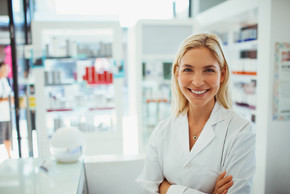 Eine lächelnde Apothekerin steht in einer Apotheke mit verschränkten Armen, im Hintergrund sind Schränke voll mit Medikamenten zu sehen.