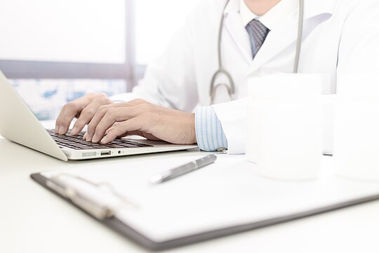  Foto: Zehn Finger auf einer Laptop-Tastatur. Tippende Person mit Stetoskop über der Schulter hängend.