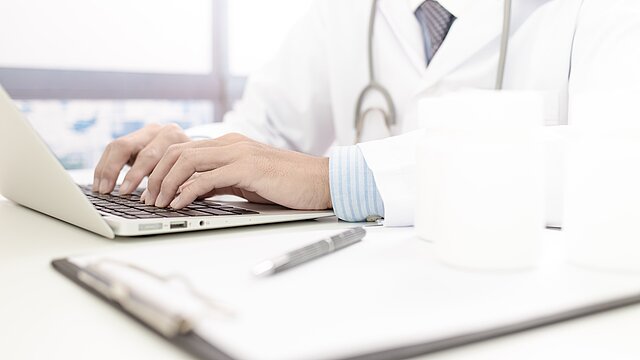  Foto: Zehn Finger auf einer Laptop-Tastatur. Tippende Person mit Stetoskop über der Schulter hängend.