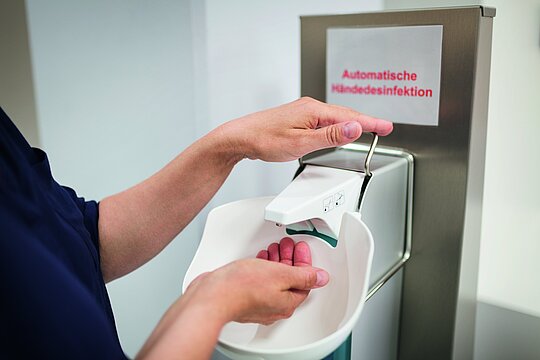 Foto: Zwei Hände einer Krankenhausmitarbeiterin an einer automatischen Händedesinfektion.