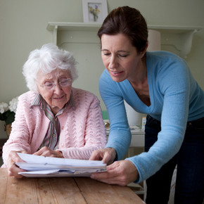 Eine Frau spricht mit einer Seniorin am Tisch über Unterlagen gebeugt