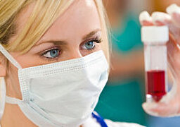Foto: Eine Frau mit Mundschutz betrachtet eine Blutprobe, die sie in der Hand auf Augenhöhe hält