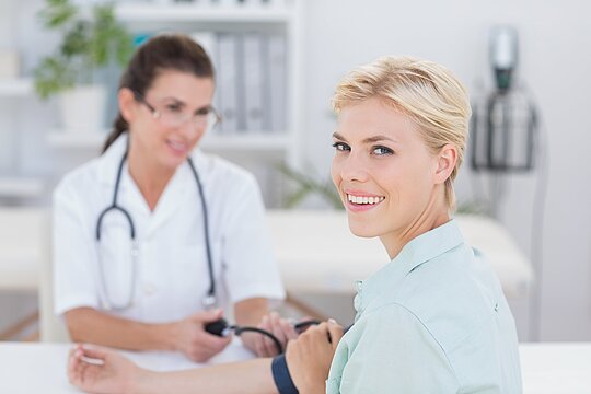 Foto: Eine dunkelhaarige Ärztin misst den Blutdruck einer blonden Patientin, die lächelnd in die Kamera schaut