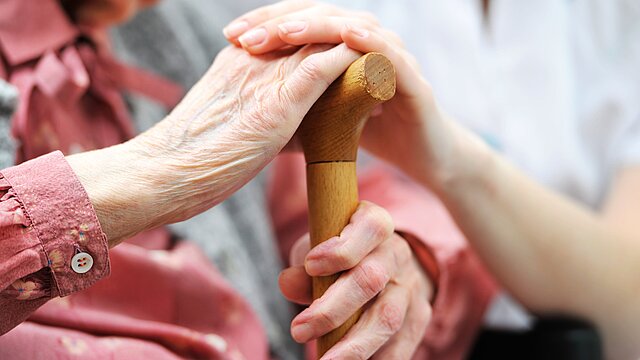 Foto: Hände einer älteren Person am Gehstock, darüber Hand einer Betreuungskraft