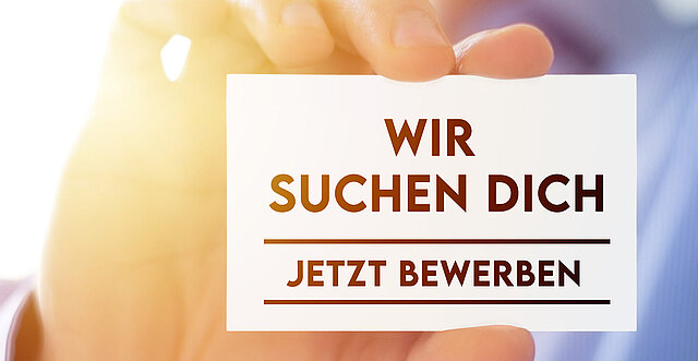 Eine Hand hält eine Karte hoch mit der Aufschrift: "Wir suchen dich - jetzt bewerben"