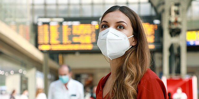 Frau mit Maske steht vor einer Bahnhofs-Anzeige
