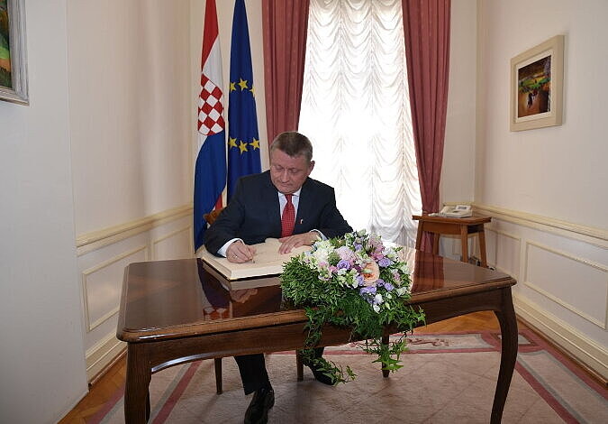 Bundesgesundheitsminister Gröhe sitzt an einem kleinen, dunklen Tisch und schreibt in ein Buch. Hinter ihm sind die kroatische und die europäische Flagge aufgestellt. Der Raum ist kaum als der Tisch, hat ein Fenster mit langen Vorhängen und es hängen Bilder an den Wänden