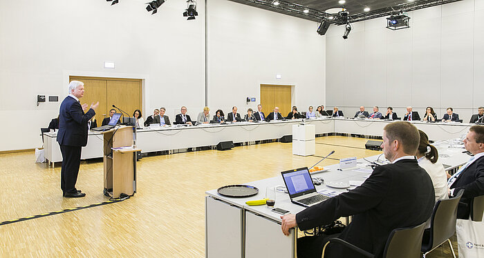 Foto: Blick in einen Workshop-Raum, ein Redner steht am Pult, im Vordergrund sind Teilnehmer zu sehen, die ihm zuhören