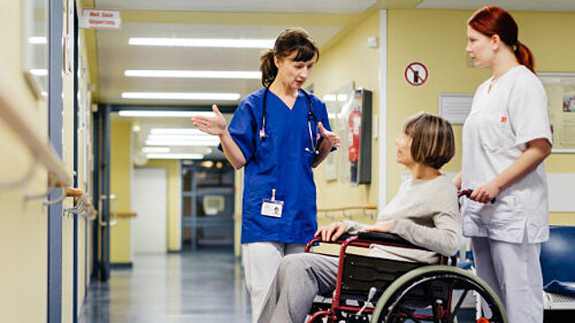 Ärztin erklärt im Krankenhausflur etwas einer älteren Patientin, die im Rollstuhl sitzt