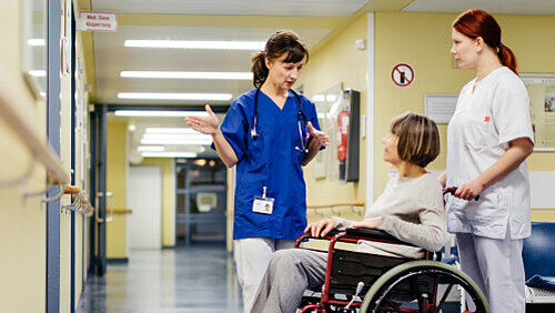 Ärztin erklärt im Krankenhausflur etwas einer älteren Patientin, die im Rollstuhl sitzt