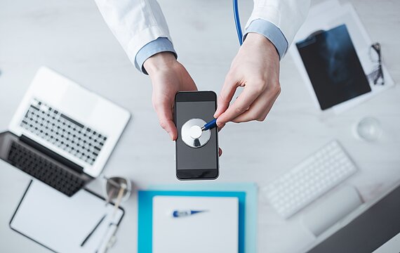 Foto: Ein Mann im Arztkittel hält ein Stethoskop an ein Mobiltelefon (Bildquelle: Shutterstock/Stokkete)