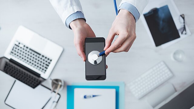 Foto: Ein Mann im Arztkittel hält ein Stethoskop an ein Mobiltelefon (Bildquelle: Shutterstock/Stokkete)