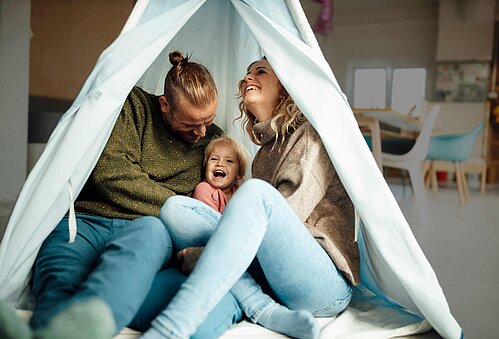 Eine 3 Köpfige Familie sitzt lachend im Wohnzimmer auf dem Boden in einem Zelt