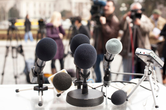 Auf einem Tisch stehen mehrere Mikrofone, im Hintergrund sind verschwommen Journalisten zu sehen. 